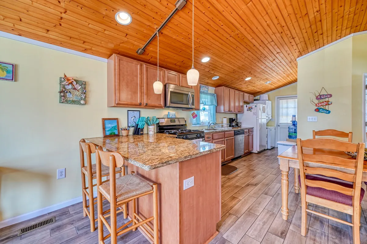 A welcoming kitchen space featuring gleaming granite countertops, modern stainless appliances, a wooden ceiling, and an adjacent dining table for four. The bright, airy ambiance encourages both meal prep and group dining, with visible counter seating creating a social hub.