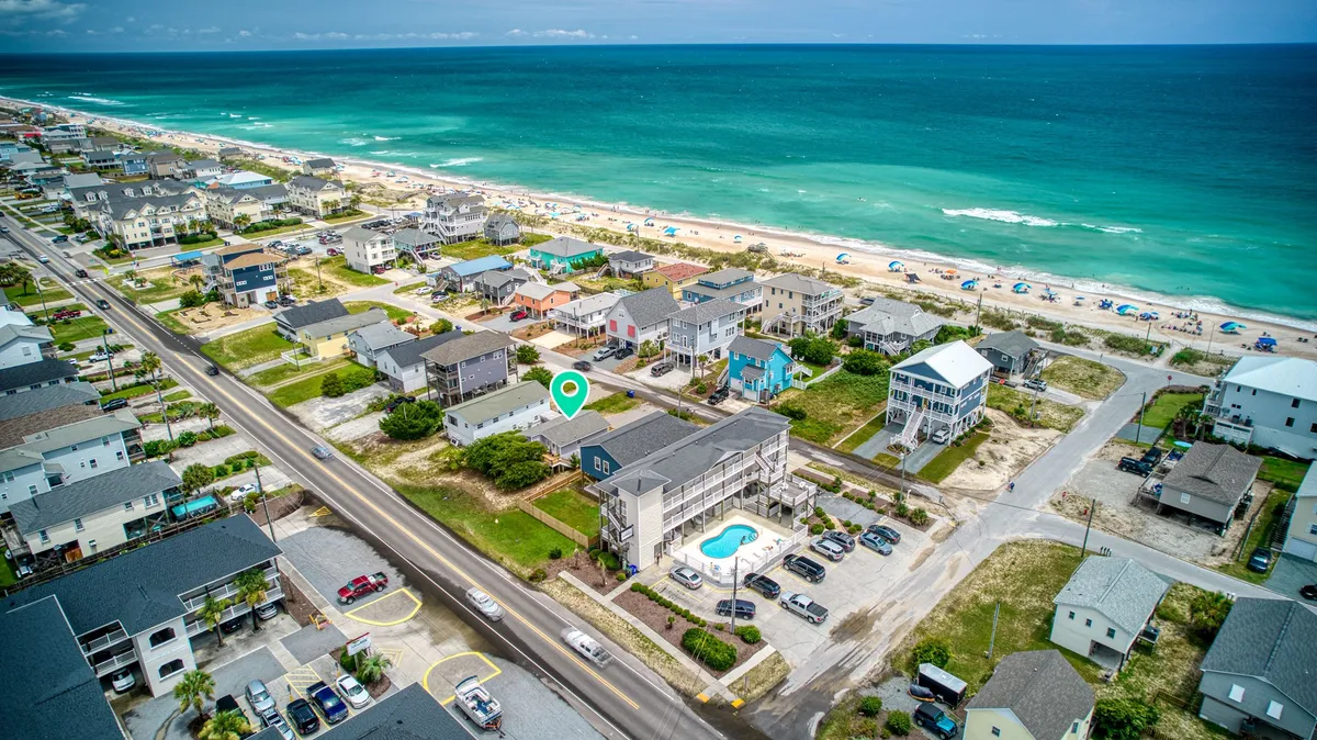 A sweeping exterior view of a row of beachfront properties lining the coast, with the sparkling ocean in full view. People are engaged in beach activities—building sandcastles, swimming, and sunbathing—creating an inviting vacation atmosphere. The image also captures the vibrant community life and easy beach access, complemented by a beautiful sunset over the water.