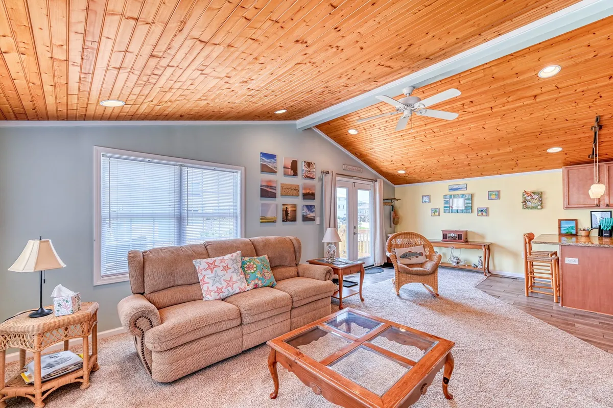 A sunlit living room with a sofa adorned with decorative pillows sits under an attractive wooden ceiling. The glass coffee table and art on the walls add a touch of personality, while open doors lead out to the porch, bathing the room in beachy natural light.