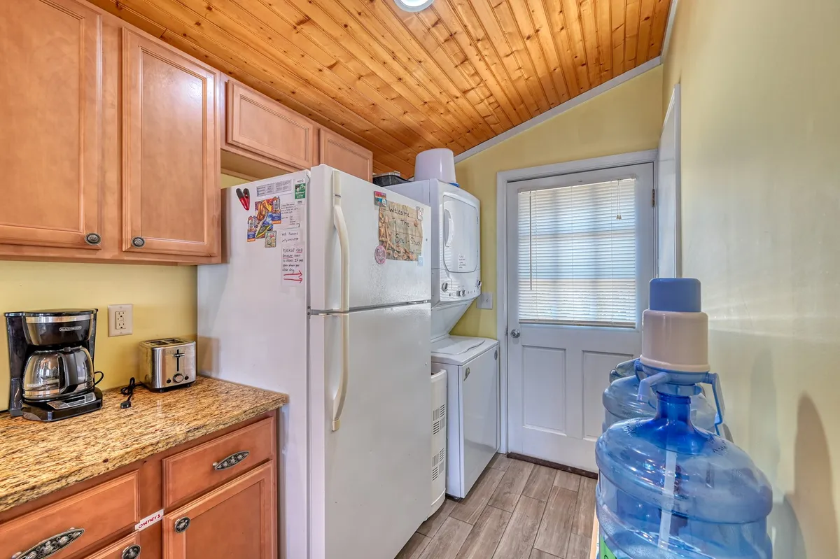 A close-up shot of the kitchen cabinetry area, showcasing a large white refrigerator, coffee maker, and a bottled water dispenser—all under soft natural light. The area is practical and welcoming, perfect for morning routines or quick refreshments.