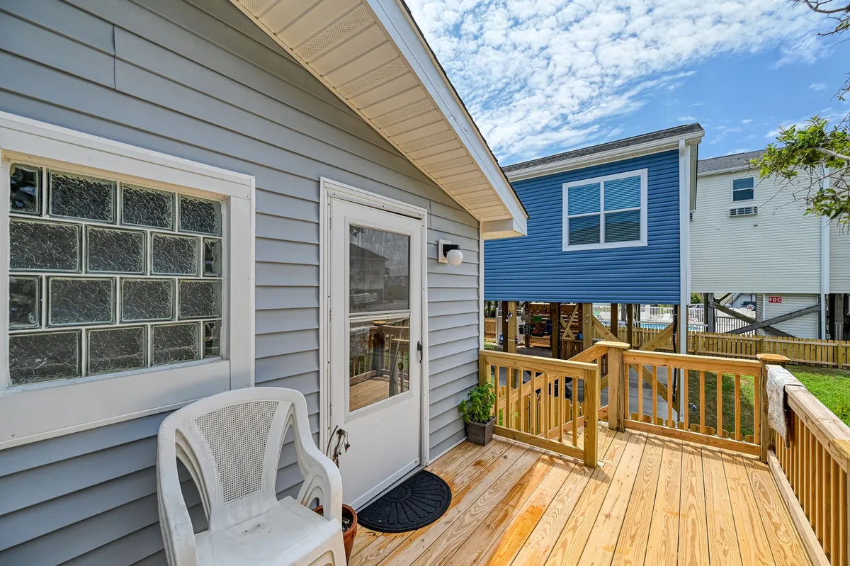 An exterior shot featuring a classic wooden deck with a glass block window and a white door, surrounded by neighboring cottages. The deck is bathed in soft sunlight, hinting at spaces for outdoor dining, grilling, and socializing after a day at the beach.