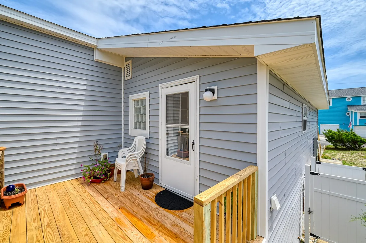 A wooden deck with a white chair, potted plants, and the front door in view. The atmosphere is inviting and tranquil, with cues for enjoying quiet moments such as sunrise coffee or reading outside. The setup is intimate and well-suited for solitary or small-group relaxation.