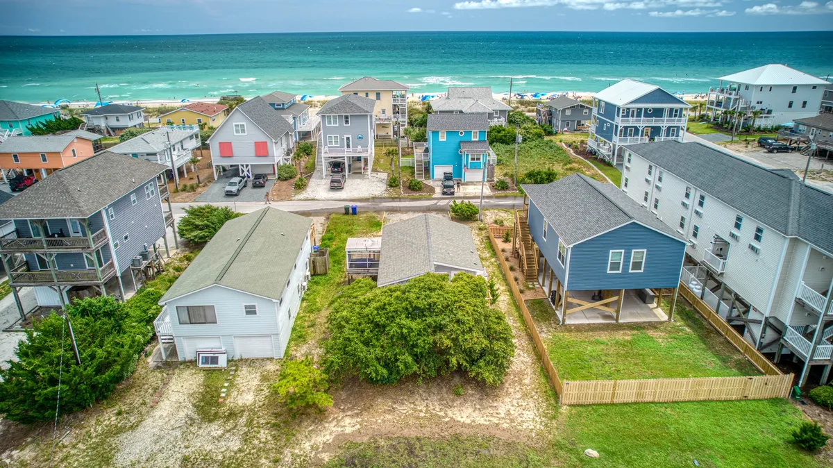A wide perspective of colorful beachfront houses, lush green vegetation, and the sandy shore, showing families gathering and enjoying both the beach and backyard spaces. The scene evokes sunset strolls, beach barbecues, and the relaxed pace of coastal life.
