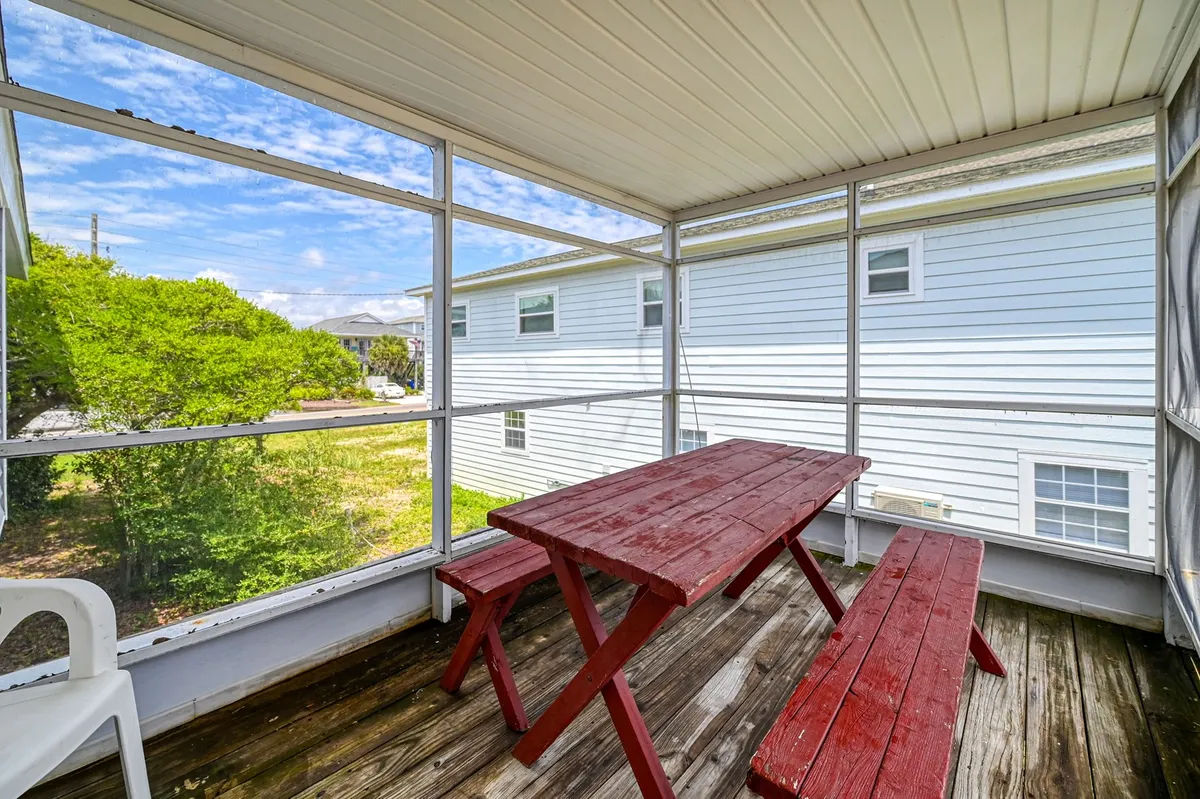 This image highlights a classic wooden picnic table set on a screened porch, with ample natural light pouring through screened windows and warm-toned wooden flooring. The setup is casual and inviting, perfect for shared meals, morning coffees, or relaxing after a day at the beach.